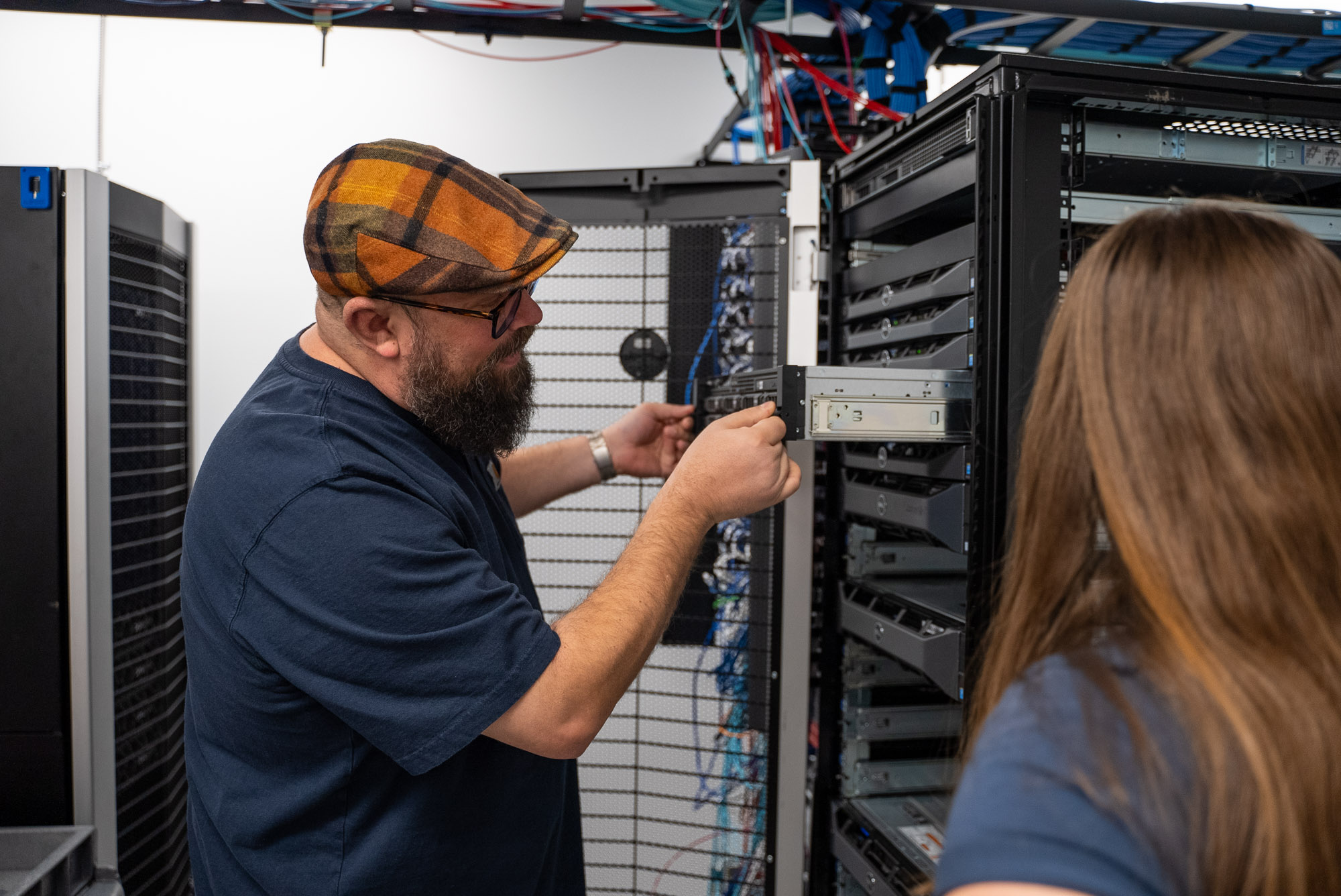 A man with a plaid hat and glasses installs a hard drive into a server rack while a woman observes.