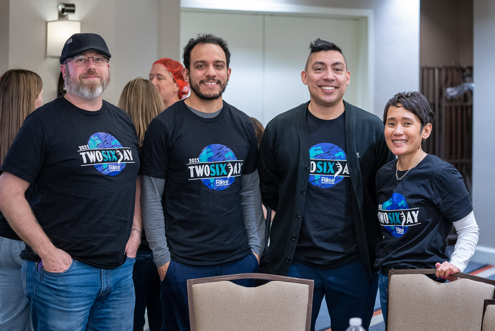 Four diverse individuals wearing "TWOSIXAY" T-shirts stand together at an indoor event, smiling for the camera.