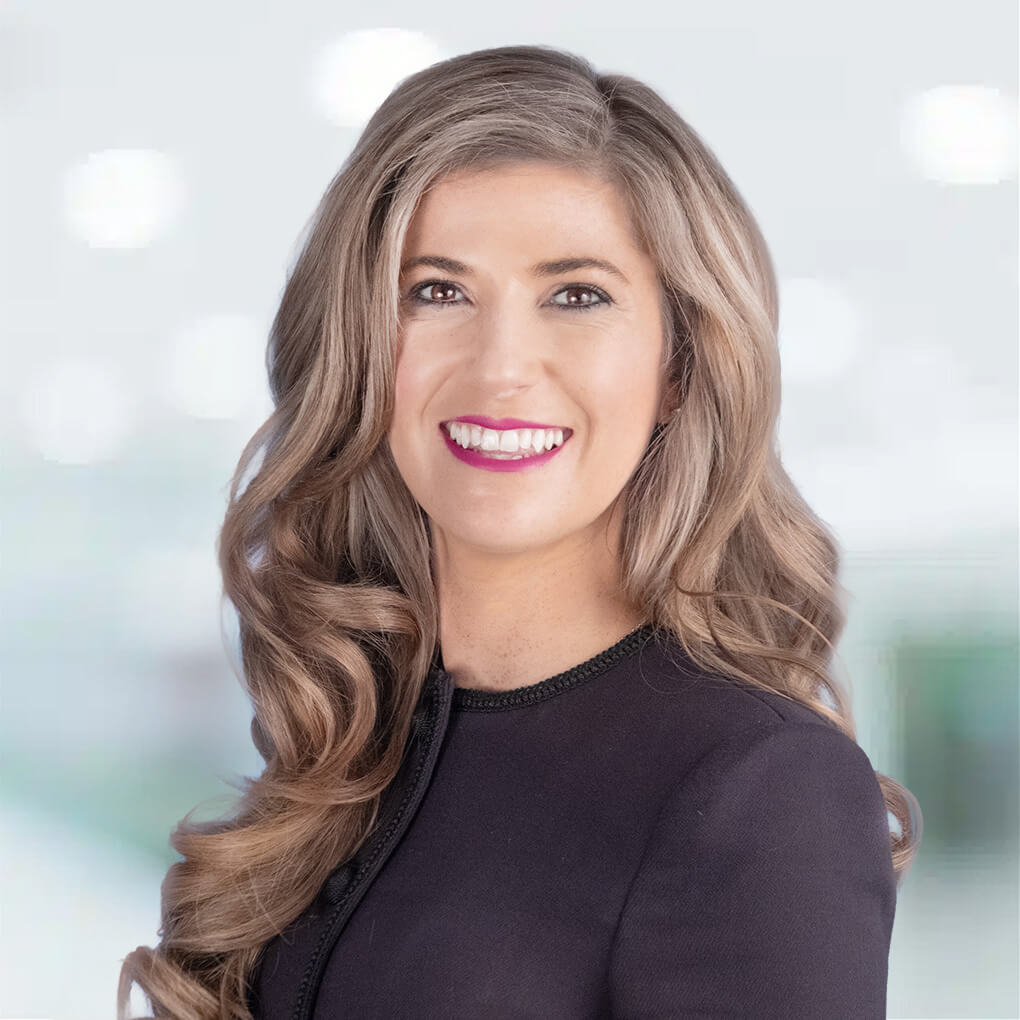 Headshot of a woman with long, wavy blonde hair, smiling with pink lipstick, wearing a black top against a blurred