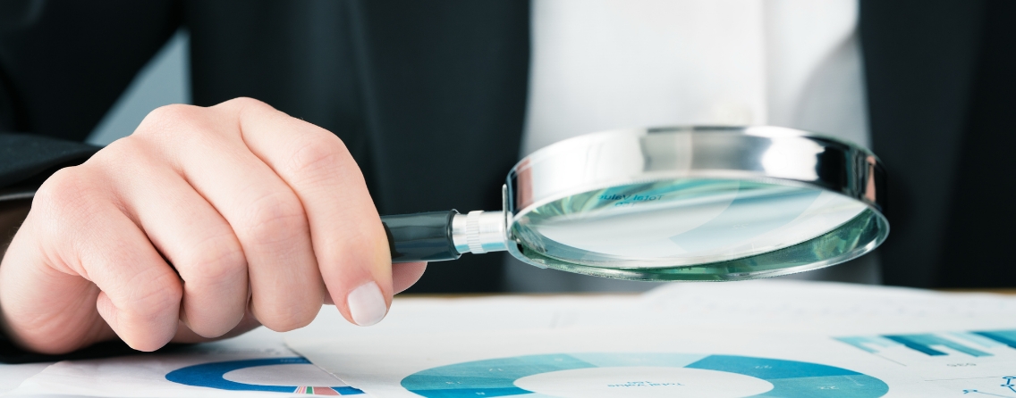 A person holds a magnifying glass over documents with charts, emphasizing auditing and monitoring processes.