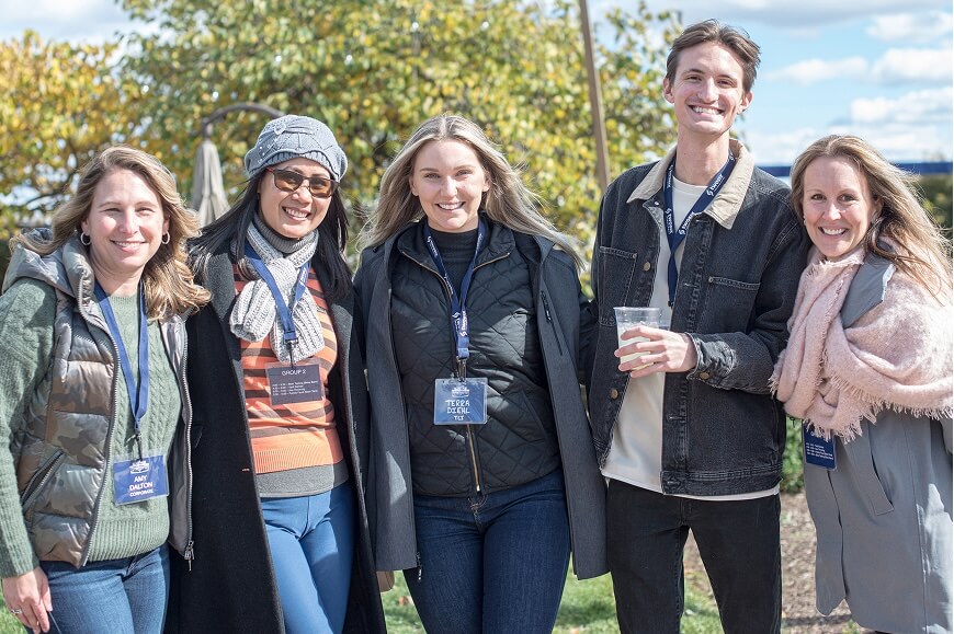 Five smiling people stand outdoors, wearing name tags and casual jackets, with trees and a blue sky in the background.