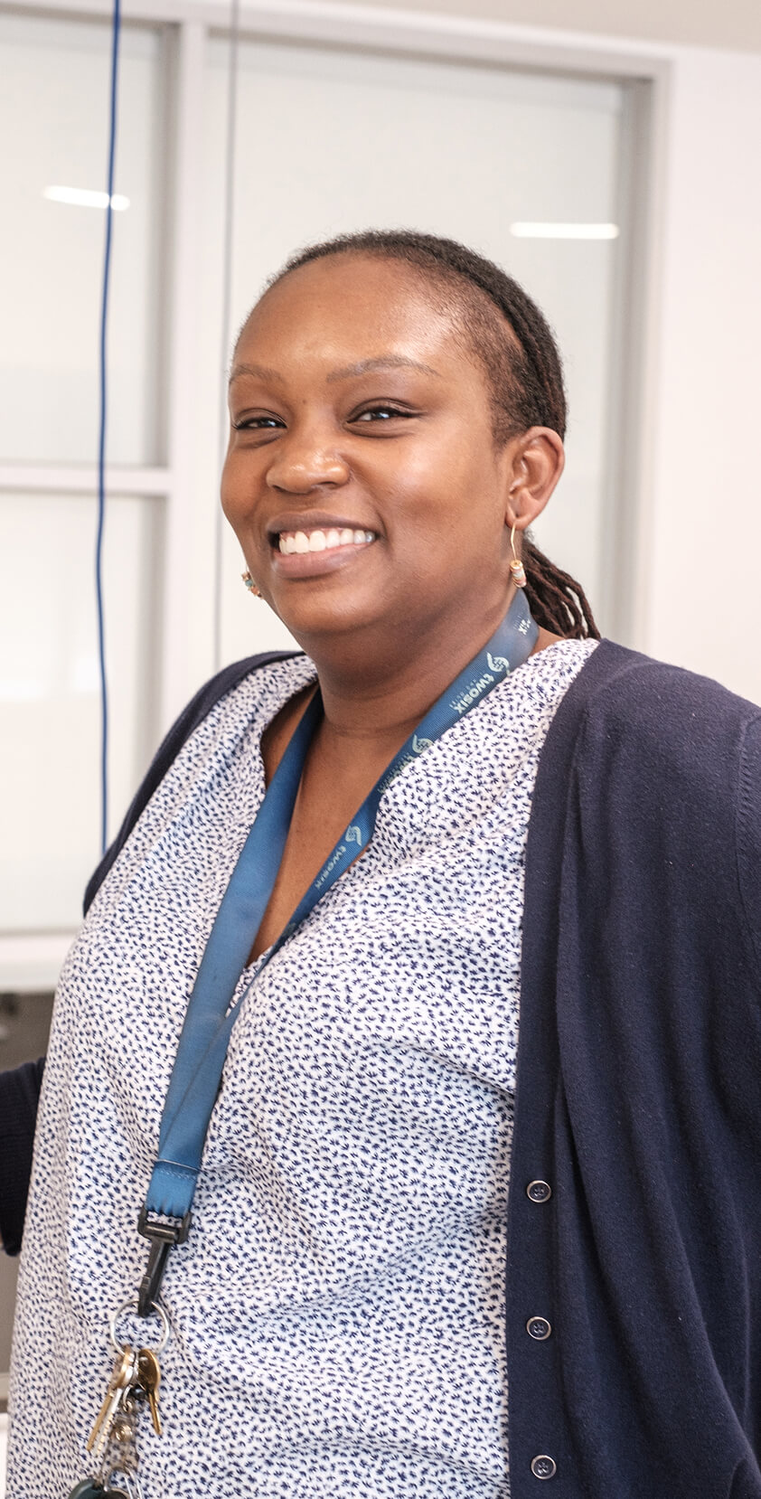 A smiling nurse with a lanyard and keys, wearing a patterned blouse and dark cardigan, standing in a clinical setting.