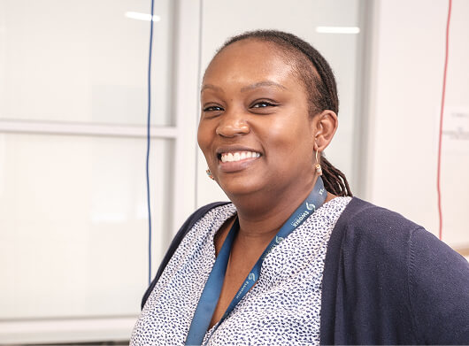 A smiling nurse with braided hair, wearing a navy cardigan and lanyard, in a clinical setting with white cabinets behind her.