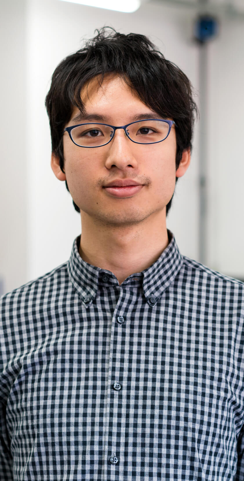 A student with black hair, glasses, and a checkered shirt, standing in a classroom setting.