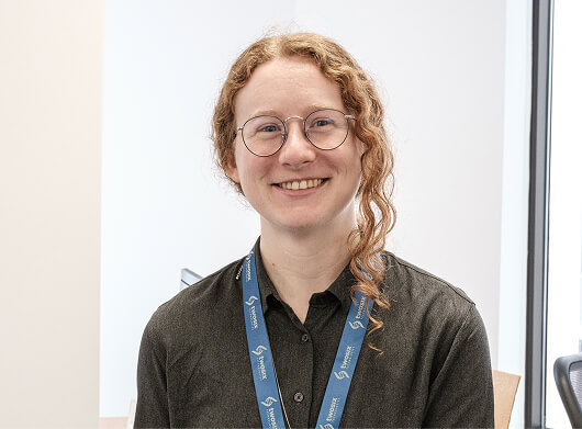 A smiling woman with curly red hair, glasses, and a lanyard, sitting in a bright office space.