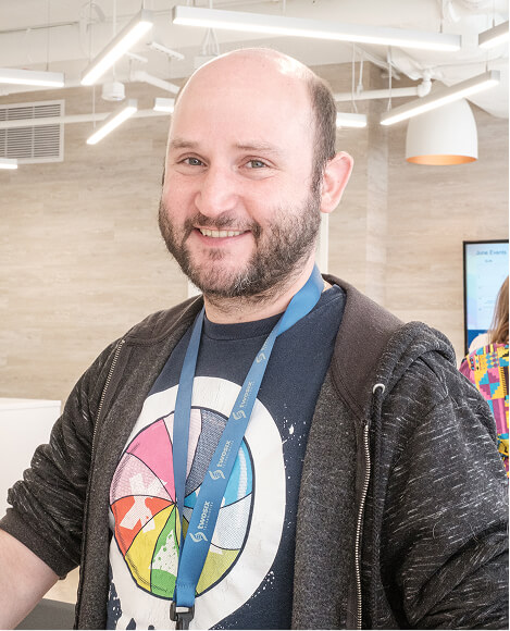 A smiling man with a beard, wearing a graphic t-shirt, black hoodie, and a blue lanyard, in a modern office space.