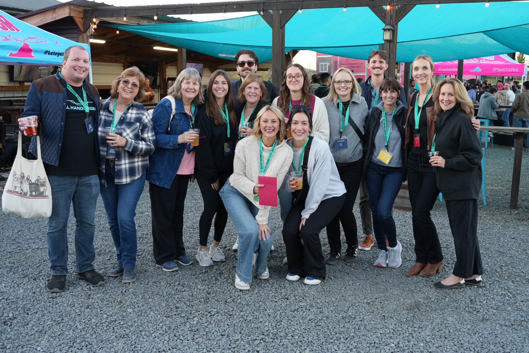 A group of smiling people wearing conference badges gather outdoors under a blue canopy, holding drinks and posing for a